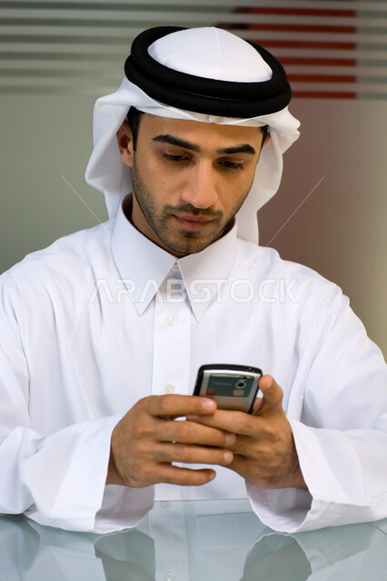 الاهتمام بالمظهر الخارجي ، صورة مقربة لشاب عربي إماراتي خليجي يرتدي الغترة و الثوب الإماراتي التقليدي ويقوم باستخام الهاتف المحمول , إنجاز مهام العمل اليومية،  تحقيق أهداف الشركة من خلال التفاهم و التعاون بين الموظفين, بيئة العمل الإماراتي . 