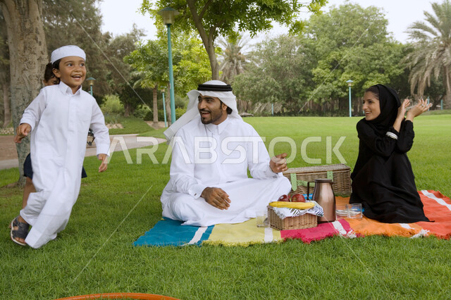 الاستمتاع في الهواء الطلق، مفهوم الترابط الأسري وحب الأبناء، قضاء أوقات ترفيهية وأنشطه ممتعه برفقه العائله، أسرة عربيه خليجيه اماراتية تقوم بالجلوس في الحديقة، النزهات والحدائق الترفيهية في الدول العربيه، أجواء عائليه مليئه بالحب.