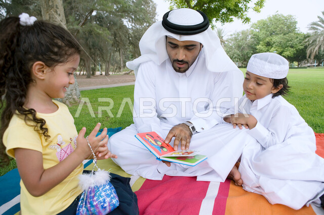 قضاء أوقات ممتعه في إحدى الحدائق في الامارات، أب عربي خليجي إماراتي يرتدي الكندوره والغتره يقوم بقراءه قصة مفيده لأولاده، مشاركه الأبناء الأنشطه الترفيهيه المفيده والمسليه، الاستمتاع في أوقات العطله مع أفراد الأسره، منتزهات وأماكن عامه في الدول العربيه.