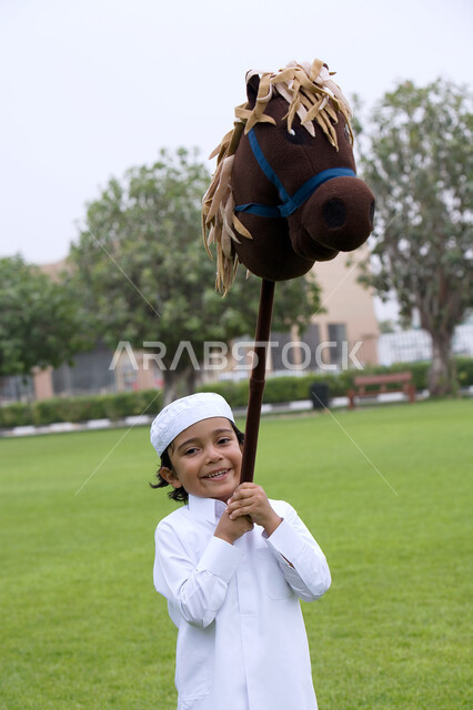 الألعاب والأنشطه الترفيهية في الحدائق العامه، ايماءات الفرح والسعاده لقضاء أوقات ممتعه في الحديقه، صبي عربي خليجي اماراتي يرتدي الزي التقليدي الاماراتي يقوم برفع لعبة الحصان، الاستمتاع في النزهة في الهواء الطلق.