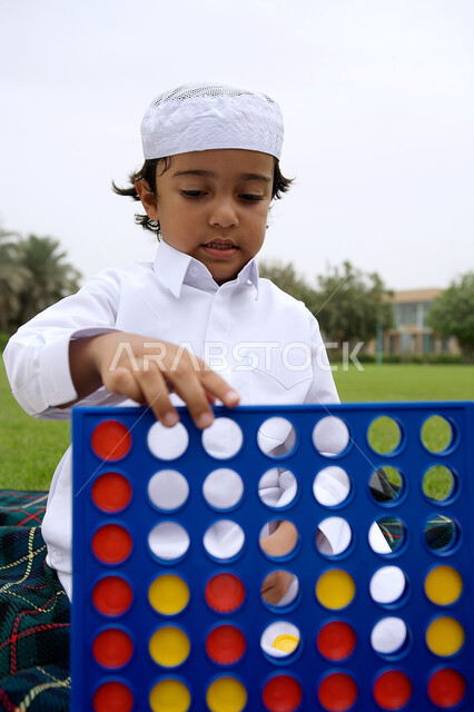 Gestures of concentration and thinking, developing intellectual and mental skills, an Arab Gulf Emirati boy wearing a traditional Emirati dress playing a four-to-win game, spending enjoyable times outdoors in one of the public parks, taking advantage of his free time by playing useful games.