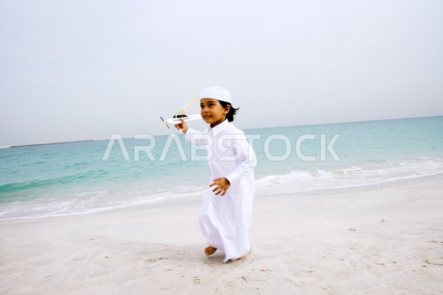 Enjoying the summer atmosphere on vacation, an Arab Gulf Emirati boy wearing a traditional costume playing a volleyball game on the seashore, entertaining recreational activities abroad, tourism in the Arab Gulf countries, beach resorts in the Emirates, entertainment and fun in free time