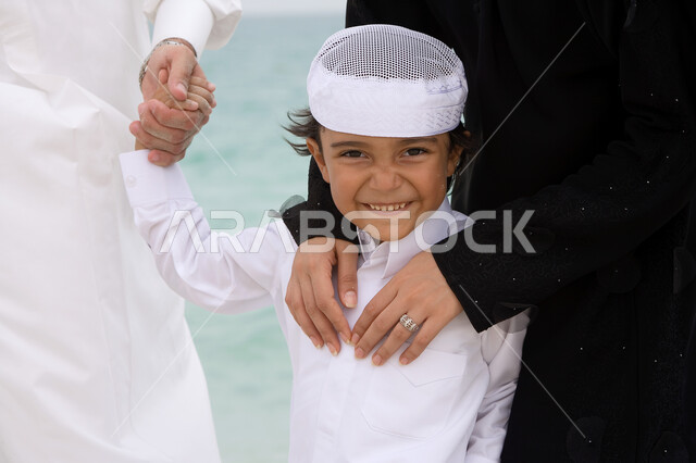  الاستمتاع في الأجواء الصيفيه على شاطئ البحر، صورة مقربة لولد عربي خليجي اماراتي يرتدي الزي التقليدي برفقه والديه ينظر إلى الكاميرا بإيماءات السعادة والسرور، قضاء أوقات الفراغ في الأماكن الساحليه مع الأولاد، السياحه في دول الخليج العربي