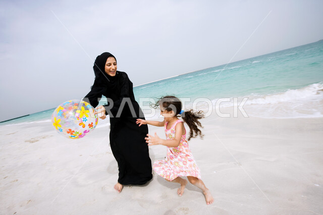 قضاء أوقات الفراغ في الأماكن الساحليه مع الأولاد, مفهوم الاسرة الإماراتية السعيدة، أم عربية خليجية إماراتية تقوم باللعب بالكرة المطاطية مع ابنتها على شاطئ البحر، السياحة في دول الخليج العربي والاستمتاع في العطلة الصيفية مع العائلة، انشطة ترفيهيه مسليه في الخارج