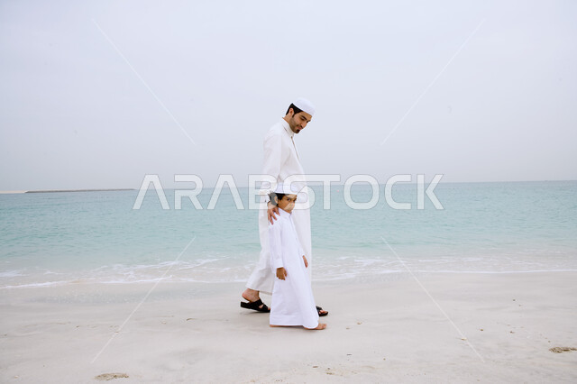 الاستمتاع بالتجول في المناطق الهادئة، صورة من الخلف لرجل عربي اماراتي خليجي يرتدي الزي التقليدي يمشي مع ولده على شاطئ البحر، نزهه في الهواء الطلق، قضاء أوقات الفراغ في الأماكن الساحليه مع الأبناء، انشطة ترفيهية مسليه، السياحة في دول الخليج العربي