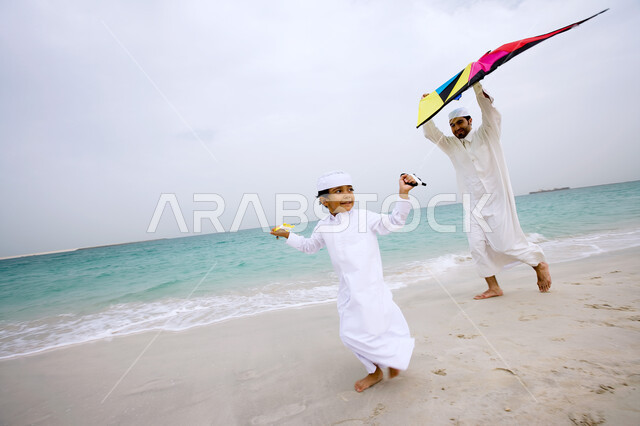 مشاركة الأبناء اللعب بالطائرة الورقيه على شاطئ البحر، أب عربي اماراتي خليجي يرتدي الزي التقليدي يقضي أوقات ممتعه مع الأولاد في الخارج، نزهه في الهواء الطلق، ايماءات تدل على السعادة والسرور، انشطة ترفيهية مسليه، السياحة في دول الخليج العربي