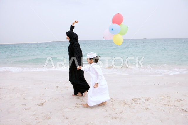 ايماءات السعاده باللعب على الشاطئ، فعاليات ترفيهية مسلية للأطفال, أم عربيه اماراتيه خليجيه محجبه ترتدي العباءه تمسك يد ابنها وتحمل بيدها مجموعه من البالونات الملونة، الاستمتاع بالأجواء الصيفيه في أوقات الفراغ مع الأولاد، التنزه في الهواء الطلق في الأماكن السياحيه