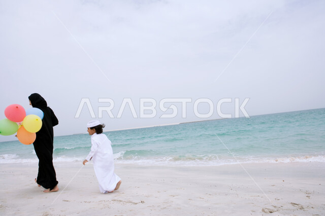 انشطة و فعاليات ترفيهية مسلية للأطفال, صورة من الخلف لأم عربيه اماراتيه محجبه ترتدي العباءه مع ابنها وتحمل بيدها مجموعه من البالونات الملونة، الاستمتاع بالأجواء الصيفيه في أوقات الفراغ مع الأولاد على الشاطئ، التنزه في الهواء الطلق في الأماكن السياحيه