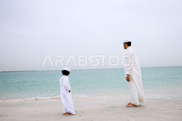 الاستمتاع بالمناظر الطبيعيه الهادئة، صورة من الخلف لرجل عربي اماراتي خليجي يرتدي الزي التقليدي يقف بجانب ولده على شاطئ البحر، نزهه في الهواء الطلق، قضاء أوقات الفراغ في الأماكن الساحليه مع الأبناء، انشطة ترفيهية مسليه، السياحة في دول الخليج العربي