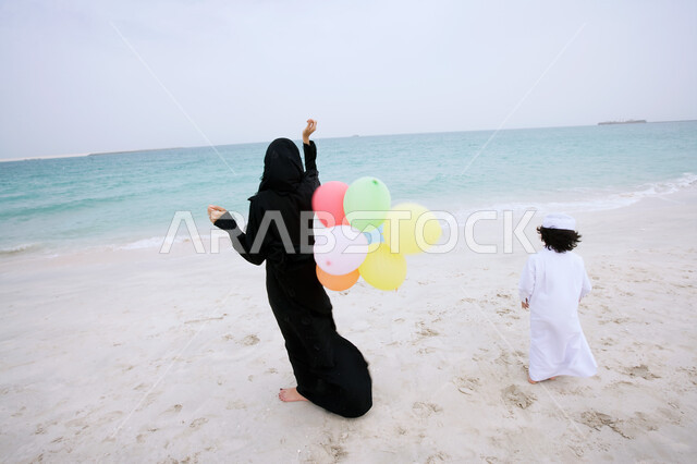 فعاليات ترفيهية مسلية للأطفال, صورة من الخلف لأم عربيه اماراتيه خليجيه محجبه ترتدي العباءه مع ابنها وتحمل بيدها مجموعه من البالونات الملونة، الاستمتاع بالأجواء الصيفيه في أوقات الفراغ مع الأولاد على الشاطئ، التنزه في الهواء الطلق في الأماكن السياحيه