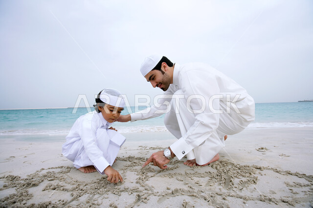 اللعب والرسم  على الرمال الذهبية الناعمة، مشاركة الآباء في النشاطات الترفيهية للأبناء, الاستمتاع بالأجواء الصيفيه على شاطئ البحر , ولد عربي خليجي اماراتي يرتدي الزي الإماراتي التقليدي يقف جانب والده بإيماءات السعاده، السياحة في دول الخليج العربي