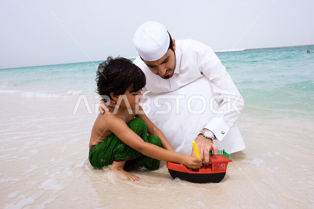 الفعاليات و الأنشطة الترفيهيه الصيفيه، مفهوم السياحه في دول الخليج العربي، ولد عربي إماراتي يقوم باللعب في الألعاب الرمليه على شاطئ البحر برفقه والده، أجواء عائليه سعيده مليئة بالحب، قضاء أوقات الفراغ في الأماكن الساحليه ،التسليه والترفيه مع الأبناء.