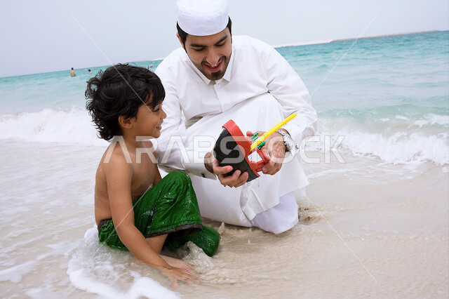 الأنشطة والفعاليات الترفيهيه الصيفيه، مفهوم السياحه في دول الخليج العربي، ولد عربي إماراتي يقوم باللعب في الألعاب الرمليه على شاطئ البحر برفقه والده، أجواء عائليه سعيده مليئة بالحب، قضاء أوقات الفراغ في الأماكن الساحليه ،التسليه والترفيه مع الأبناء.