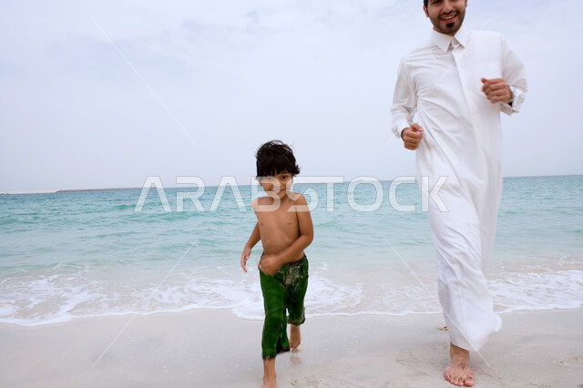 الركض على رمال البحر الناعمة، انشطة ترفيهية ممتعه، مفهوم الاسرة الاماراتيه السعيدة، رجل اماراتي خليجي يرتدي الزي التقليدي يلعب مع ولده على الشاطئ، نزهه في الهواء الطلق، قضاء أوقات الفراغ في الأماكن الساحليه مع الأبناء، السياحة في دول الخليج العربي