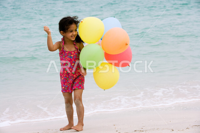 أنشطه ترفيهية مسلية للأطفال, الاستمتاع بالأجواء الصيفيه على شاطئ البحر، فتاة إماراتية ترتدي ملابس السباحة وتحمل بيدها مجموعه من البالونات الملونة, السياحة في دول الخليج العربي، قضاء أوقات الفراغ في الأماكن الساحليه، صورة الجسم بالطول الكامل