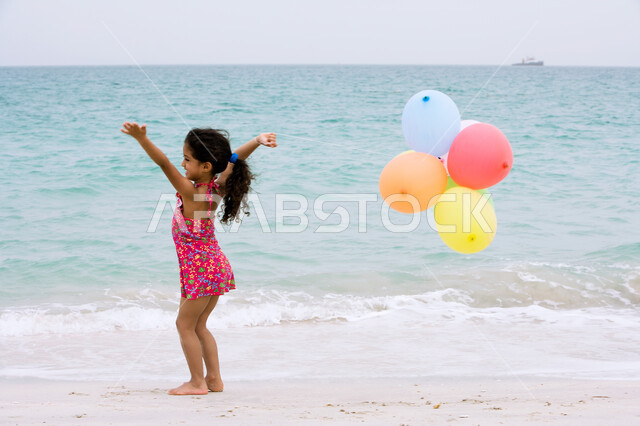 الاستمتاع بالأجواء الصيفيه على شاطئ البحر، التنزه في الهواء الطلق, أنشطه ترفيهية مسلية للأطفال, صورة من الخلف لفتاة إماراتية ترتدي ملابس السباحة وتحمل بيدها مجموعه من البالونات الملونة, السياحة في دول الخليج العربي، قضاء أوقات الفراغ في الأماكن الساحليه