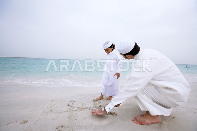 الرسم و اللعب بالرمال الذهبية الناعمة، مشاركة الآباء في النشاطات الترفيهية للأبناء, الاستمتاع بالأجواء الصيفيه على شاطئ البحر , ولد عربي خليجي اماراتي يرتدي الزي الإماراتي التقليدي يقف جانب والده بإيماءات السعاده، السياحة في دول الخليج العربي