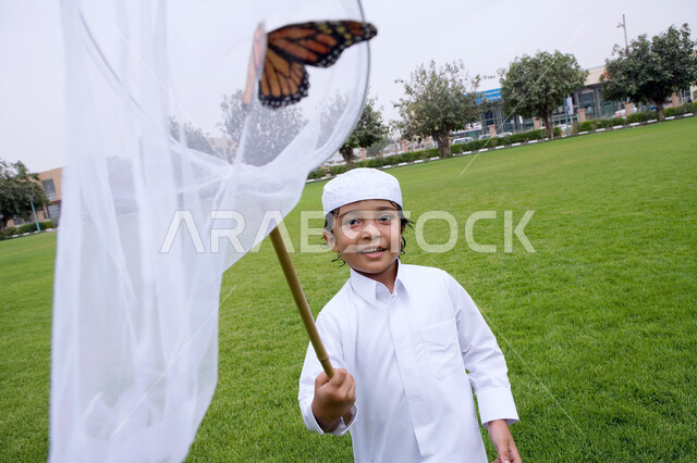 قضاء أوقات ممتعه في الحدائق العامه، أنشطه ترفيهية مسليه للأطفال، الاستمتاع بالركض في الطبيعة الخضراء في الهواء الطلق، صبي عربي خليجي اماراتي يرتدي الثوب الاماراتي التقليدي يقوم بإصطياد الفراشات بواسطة شبكة الصيد، ايماءات الفرح والسعاده.
