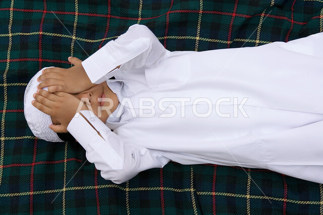 الشعور بالإحراج والإنعزال عن الناس، صبي عربي خليجي اماراتي يرتدي الثوب التقليدي الاماراتي يستلقي على الحصيره ويقوم بتخبآة وجهه بواسطه يديه، مفهوم الرهاب الإجتماعي لدى الأطفال، ايماءات تدل على الخجل.