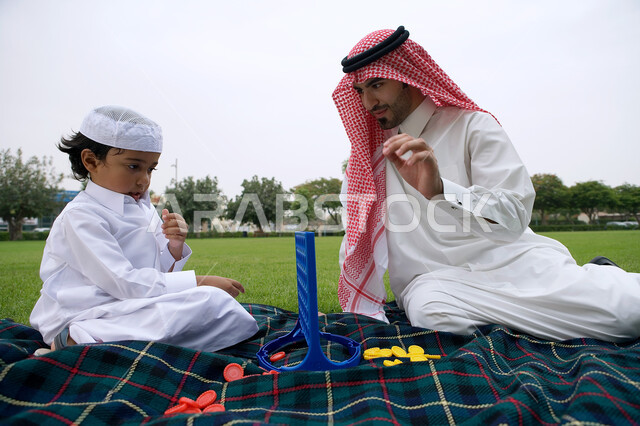 ايماءات التركيز والتفكير ،فضاء أوقات ممتعة في الهواء الطلق في إحدى الحدائق العامه، الاستفاده من أوقات الفراغ في اكتساب القدرات الفكرية، تنمية المهارات العقليه والذهنيه، أب عربي خليجي اماراتي يرتدي الزي الاماراتي التقليدي يتنافس مع ابنه بلعبه اربع تربح .