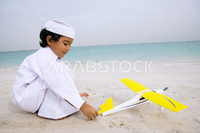  التسليه والمرح في أوقات الفراغ، الاستمتاع بالأجواء الصيفيه في العطلة، ولد عربي خليجي اماراتي يرتدي الزي التقليدي يقوم باللعب بلعبة الطائرة على شاطئ البحر، انشطة ترفيهيه مسليه في الخارج، السياحة في دول الخليج العربي ، منتجعات شاطئية في الإمارات
