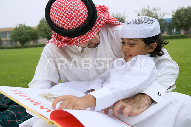 أماكن و منتزهات عامه في دول الخليج العربي، مشاركه الأبناء الأنشطه الترفيهيه والمسليه، قضاء أوقات ممتعه في إحدى الحدائق ، صورة مقربة لرجل عربي خليجي إماراتي يرتدي الزي التقليدي يقوم بقراءه قصة مفيده لابنه، الاستمتاع في أوقات العطله مع أفراد الأسره