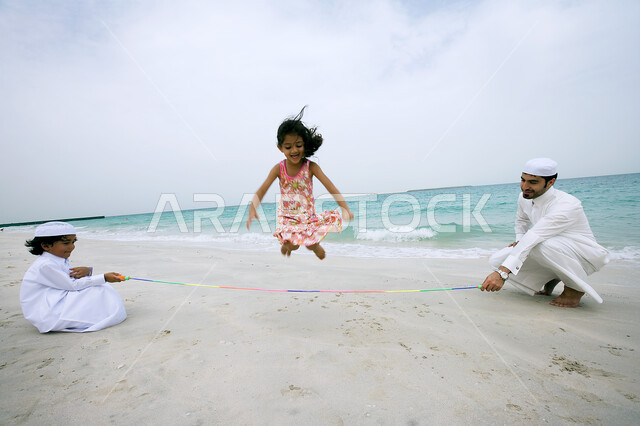 ممارسة لعبه القفز على الحبل فوق الرمال الناعمة، قضاء أوقات الفراغ في الأماكن الساحليه، رجل عربي خليجي إماراتي يقوم بمشاركة أولاده اللعب على شاطئ البحر، الاستمتاع بالأجواء الصيفيه بإيماءات السعاده والسرور ،السياحه في دول الخليج العربي