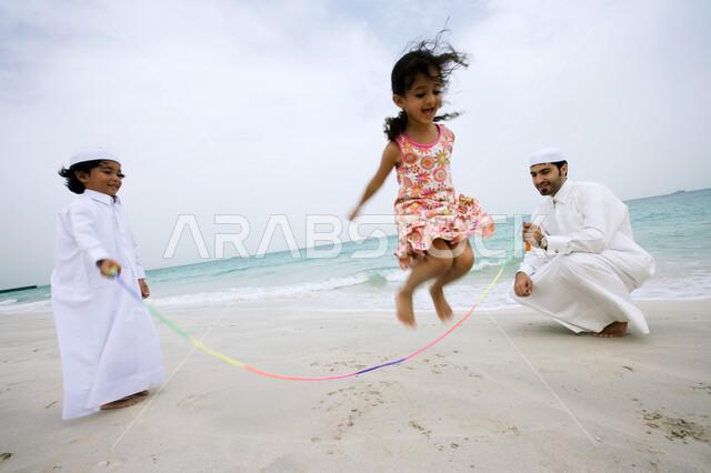 Enjoying the summer atmosphere with gestures of happiness and pleasure, playing the game of jumping rope over the soft sand, spending leisure time in coastal places, an Arab Gulf Emirati man who shares with his children playing on the seashore, tourism in the Arab Gulf countries