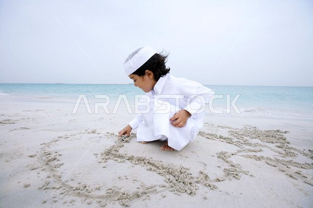 Entertaining events and activities, enjoying the summer atmosphere on the seashore, an Emirati Arab boy wearing the traditional Emirati costume playing and drawing on the soft golden sands, spending leisure time in coastal places, tourism in the Arab Gulf countries