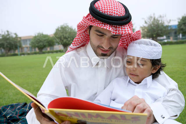 منتزهات وأماكن عامه في دول الخليج العربي، مشاركه الأبناء الأنشطه الترفيهيه والمسليه، قضاء أوقات ممتعه في إحدى الحدائق ، صورة مقربة لرجل عربي خليجي إماراتي يرتدي الزي التقليدي يقوم بقراءه قصة مفيده لابنه، الاستمتاع في أوقات العطله مع أفراد الأسره