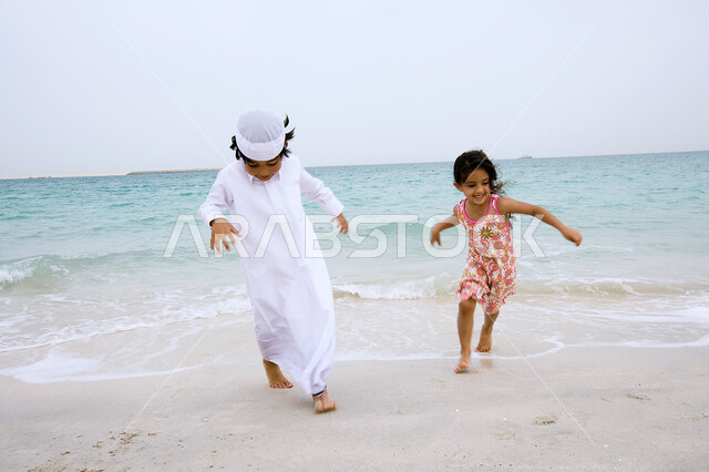 اللعب والركض على الرمال بإيماءات السعاده، الإستمتاع في الفعاليات الصيفية في الخارج, فتاة صغيرة عربية إماراتية تقوم باللعب على شاطئ البحر رفقة أخيها , منتجعات شاطئية في الإمارات العربيه المتحده، قضاء أوقات الفراغ  في الأماكن الساحليه في دول الخليج العربي