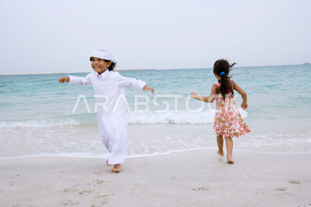 Running and playing in the sand with gestures of happiness, enjoying summer activities outside, a young Arab Emirati girl playing on the seashore with her brother, beach resorts in the United Arab Emirates, spending leisure time in coastal places in the Arab Gulf countries