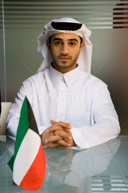 الاهتمام بالمظهر الخارجي و الأناقة ، صورة مقربة لموظف عربي إماراتي خليجي يرتدي الغترة و الثوب الإماراتي التقليدي وينظر إلى الكاميرا  ,إيماءات وجه تدل على الثقة بالنفس والطموح  , علم الإمارات العربية المتحدة  ، خلفية جدار زجاجي . 
