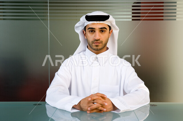 الأناقة و الاهتمام بالمظهر الخارجي ، صورة مقربة لموظف عربي إماراتي خليجي يرتدي الغترة و الثوب الإماراتي التقليدي وينظر إلى الكاميرا  ,إيماءات وجه تدل على الثقة بالنفس والطموح  , بيئة العمل في الإمارات  ، خلفية جدار زجاجي . 