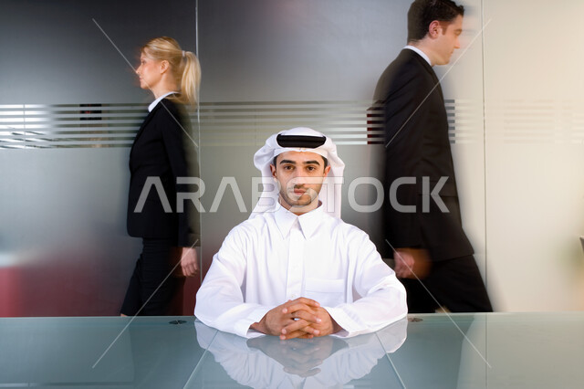تحركات وتنقل رجال الأعمال والموظفين في مقر الشركة  ، رجل عربي إماراتي خليجي يرتدي الغترة و الثوب الإماراتي التقليدي ويينظر إلى الكاميرا  ,إيماءات وجه تدل على الثقة بالنفس والطموح  , بيئة العمل في الإمارات  ، خلفية جدار زجاجي . 