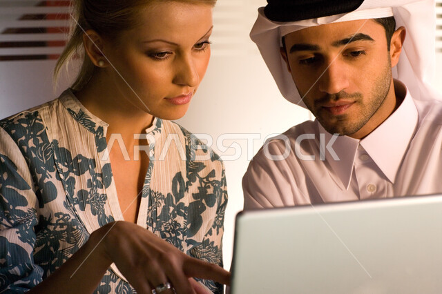  مناقشة المهام  والاستراتيجيات  ، رجل أعمال عربي إماراتي خليجي يرتدي الغترة و الثوب الإماراتي التقليدي يجلس بجانب سيدة و يقومون بالتعاون لتحقيق أهداف الشركة ,  إستخدام جهاز الحاسوب المحمول في إنجاز الأعمال ،  بيئة العمل في  الإمارات ، خلفية جدار زجاجي  .