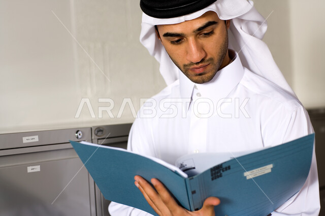  تحقيق أهداف الشركة بكفاءة عالية  ، صورة مقربة لشاب عربي إماراتي خليجي يرتدي الغترة و الثوب الإماراتي التقليدي ويبحث في الملفات التعريفية , إنجاز مهام العمل اليومية، الأناقة والاهتمام بالمظهر الخارجي ، إيماءات وجه تدل على القراءة ، بيئة العمل الإماراتي . 