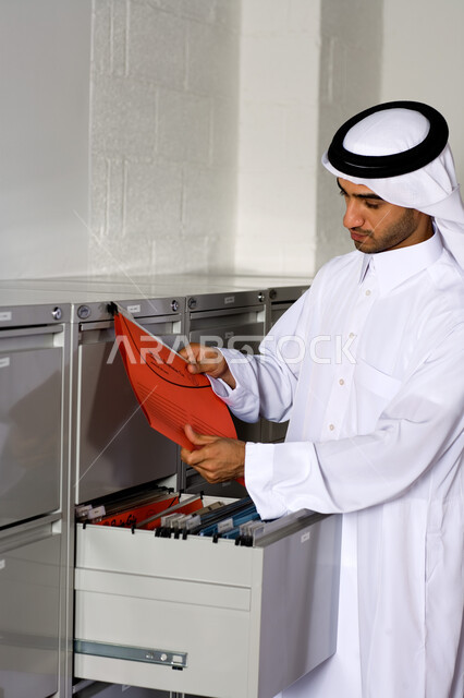 التأكد والتحقق من الملفات  ، صورة مقربة لشاب عربي إماراتي خليجي يرتدي الغترة و الثوب الإماراتي التقليدي ويبحث في الملفات التعريفية , إنجاز مهام العمل اليومية،  تحقيق أهداف الشركة بكفاءة عالية ، الأناقة والاهتمام بالمظهر الخارجي ،  بيئة العمل في الإمارات . 