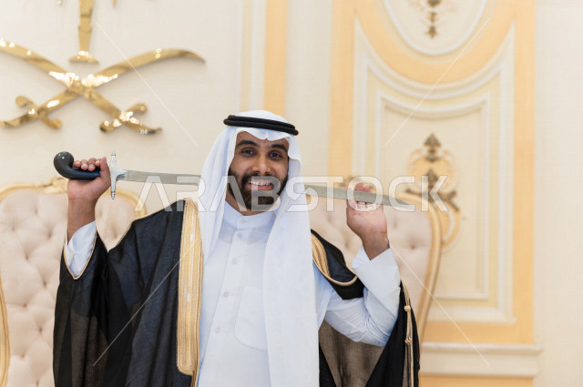 معرس شاب سعودي خليجي بتعابير وجه تدل على السعادة ،  يرقصو يحمل بيده السيف ، عريس بالزي السعودي الرسمي ، مناسبات الزواج