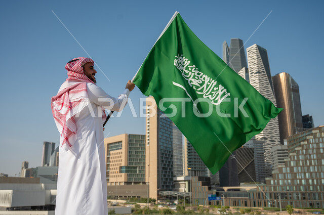 الفن الهندسي المعماري في أبراج وناطحات السحاب في الرياض، صورة من الخلف لرجل عربي خليجي سعودي يرتدي الزي التقليدي ويرفع علم السعودية، ذكرى اليوم الوطني السعودي، يوم العلم السعودي 11 مارس، العيد الوطني 23 سبتمبر، خلفية مركز الملك عبد الله المالي كافد