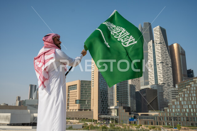 النمور والتطور العمراني في المباني والمنشآت المالية في الرياض، صورة من الخلف لرجل عربي خليجي سعودي يرتدي الزي التقليدي ويرفع علم السعودية، اليوم الوطني السعودي، يوم العلم السعودي 11 مارس، العيد الوطني 8 ربيع الأول، خلفية مركز الملك عبد الله المالي (كافد)