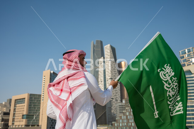 الفن الهندسي المعماري لمركز الملك عبد الله المالي (كافد)، صورة من الخلف لرجل عربي خليجي سعودي يرتدي الزي التقليدي ويرفع علم السعودية، اليوم الوطني السعودي، يوم العلم السعودي 11 مارس، العيد الوطني 23 سبتمبر، خلفية المباني والمنشآت المالية في مدينة الرياض