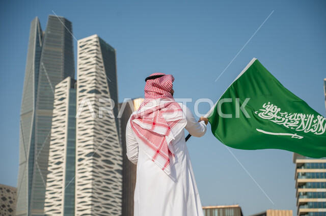 يوم العلم السعودي 11 مارس، صورة من الخلف لرجل عربي خليجي سعودي يرتدي الزي التقليدي ويرفع علم السعودية، ذكرى اليوم الوطني 23 سبتمبر، العيد الوطني السعودي، النمو والتطور العمراني في الأبراج وناطحات السحاب في مدينة الرياض، خلفية مركز الملك عبد الله المالي
