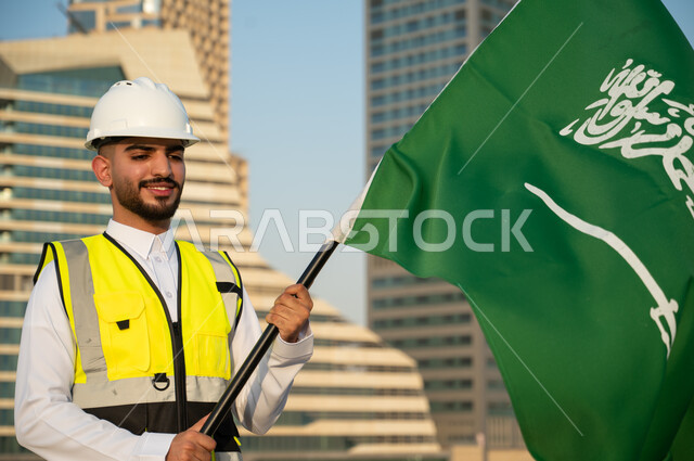 يوم العلم السعودي 11 مارس، مفهوم الإنشاءات والهندسة المعمارية، مهندس عربي خليجي سعودي يرتدي الخوذة وسترة الحماية ويحمل علم السعودية بسعادة وفخر، اليوم الوطني السعودي، العيد الوطني السعودي 23 سبتمبر، خلفية مركز الملك عبد الله المالي في الرياض