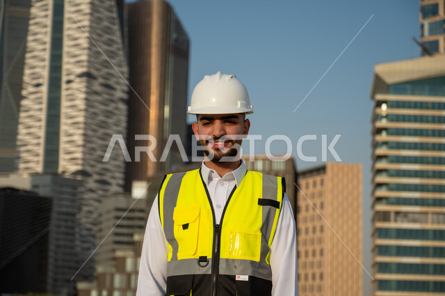 التطور العمراني بسواعد أبناء الوطن، مهندس عربي خليجي سعودي مبتسم يرتدي الخوذة وسترة الحماية، الإنشاءات والهندسة المعمارية، المشاريع الهندسية في السعودية، العمل في القطاع الهندسي السعودي، أبراج مالية ومنشآت اقتصادية في الرياض، خلفية مركز الملك عبدالله المالي