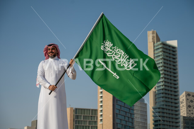 مشاعر الاعتزاز بالهوية الوطنية، النمور والتطور العمراني في مدينة الرياض، رجل عربي خليجي سعودي يرتدي الزي التقليدي ويرفع علم السعودية بسعادة وثقة، اليوم الوطني السعودي، يوم العلم السعودي 11 مارس، العيد الوطني 8 ربيع الأول، خلفية مركز الملك عبد الله المالي