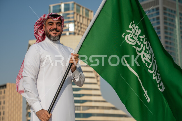 مشاعر الحب والولاء للمملكة، النمور والتطور العمراني في مدينة الرياض، رجل عربي خليجي سعودي يرتدي الزي التقليدي ويرفع علم السعودية بسعادة وثقة، اليوم الوطني السعودي، يوم العلم السعودي 11 مارس، العيد الوطني 8 ربيع الأول، خلفية مركز الملك عبد الله المالي