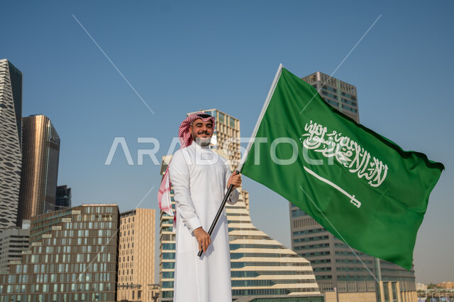 الفخر بالهوية الوطنية السعودية، النمو والتطور العمراني في مدينة الرياض، رجل عربي خليجي سعودي يرتدي الزي التقليدي ويرفع علم السعودية بسعادة وثقة، اليوم الوطني السعودي، يوم العلم السعودي 11 مارس، العيد الوطني 8 ربيع الأول، خلفية مركز الملك عبد الله المالي