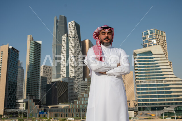الوقوف بثقة والنظر الى الكاميرا، النمو والتطور العمراني في السعودية، رجل عربي خليجي سعودي يرتدي الشماغ والثوب التقليدي بايدي مكتوفة، الفن الهندسي المعماري لمركز الملك عبد الله المالي (كافد)، معالم الرياض المشهورة، خلفية الأبراج وناطحات السحاب في الرياض