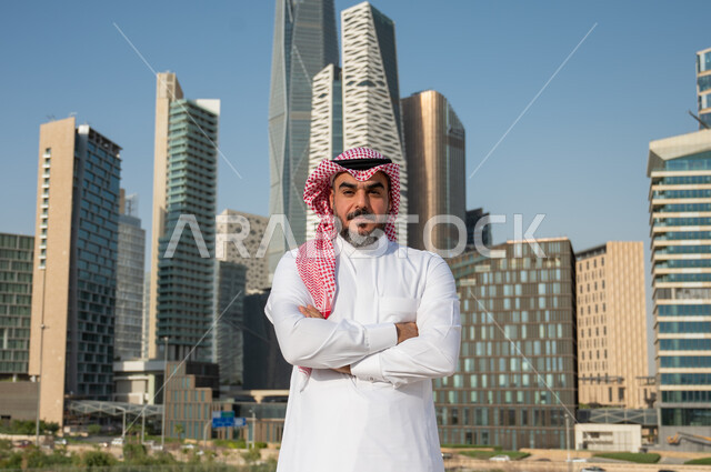 الفن الهندسي المعماري لمركز الملك عبد الله المالي (كافد)، رجل عربي خليجي سعودي يرتدي الشماغ والثوب التقليدي بايدي مكتوفة، الوقوف بثقة والنظر الى الكاميرا، النمو والتطور العمراني في السعودية، معالم الرياض المشهورة، خلفية الأبراج وناطحات السحاب في الرياض